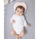 Baby lying on a gray surface, wearing a white onesie with colorful dots, with a pink Babymoov Lovenest pillow shaped like a heart behind their head.