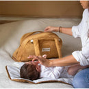 Bbluv camel-colored changing bag next to a baby on a changing mat, with a caregiver reaching for the bag.