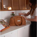 Doudoune Changing Bag by Bbluv in camel color, being opened by a woman at a countertop, with a rose in background.