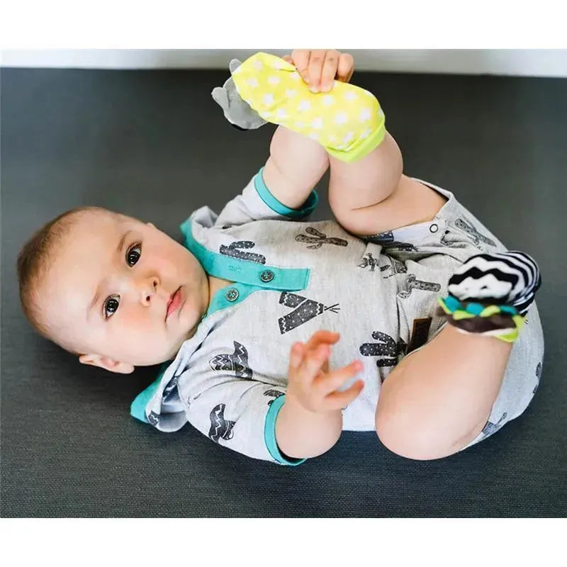 Baby wearing Bbluv Duo Foot Finders, featuring a soft yellow foot cover with a hedgehog and a black and white striped cover with a raccoon, playing on a gray surface.