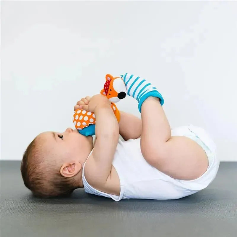 Baby playing on a gray surface, holding Bbluv Duo Foot Finders in fox and owl designs, wearing a white onesie.