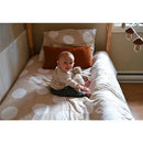Infant sitting on a bed with polka dot bedding, smiling and holding a stuffed animal, with a decorative pillow nearby.