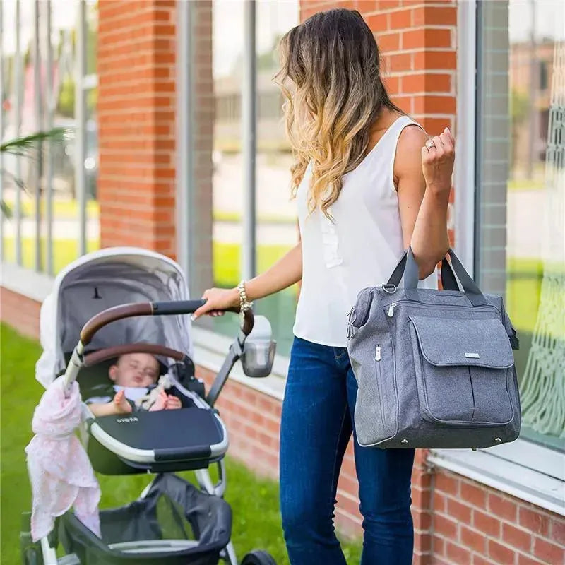 Charcoal Bbluv Metrö Complete Backpack Diaper Bag held by a woman beside a stroller with a sleeping baby.