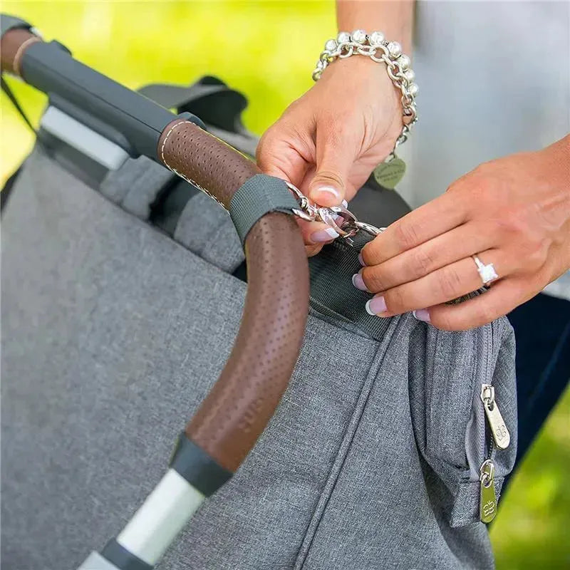 User attaching a Bbluv Metrö Complete Backpack Diaper Bag, charcoal gray, to a stroller with a clip.