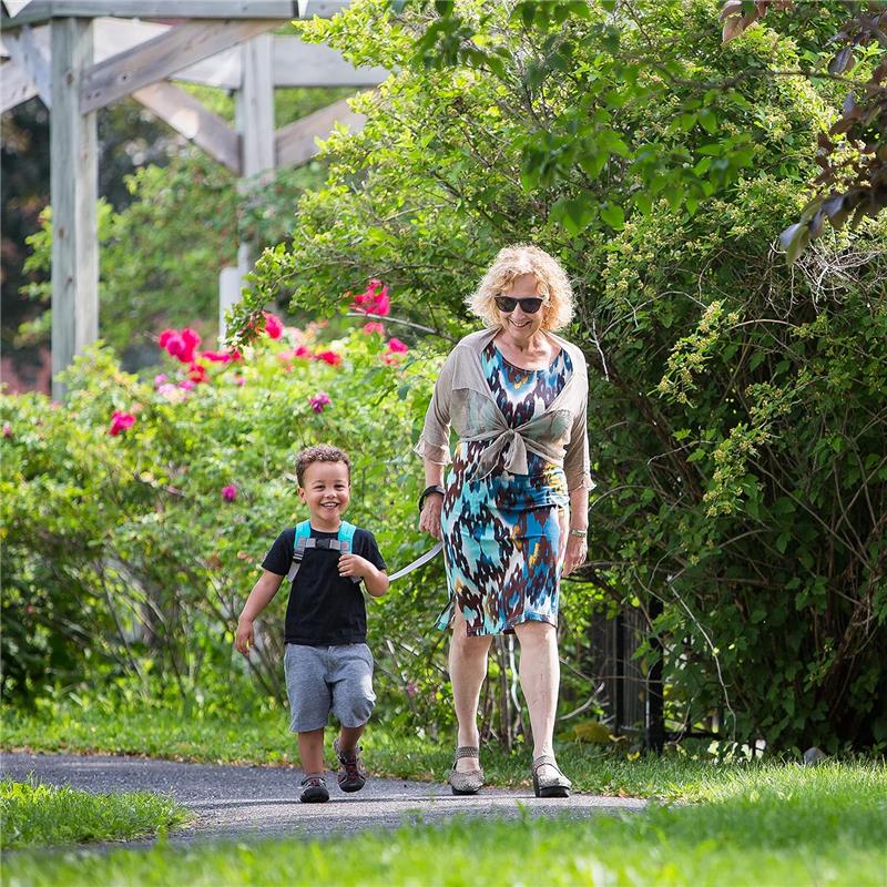 A child wearing a toddler backpack with rein joyfully walks beside an adult in a garden setting.