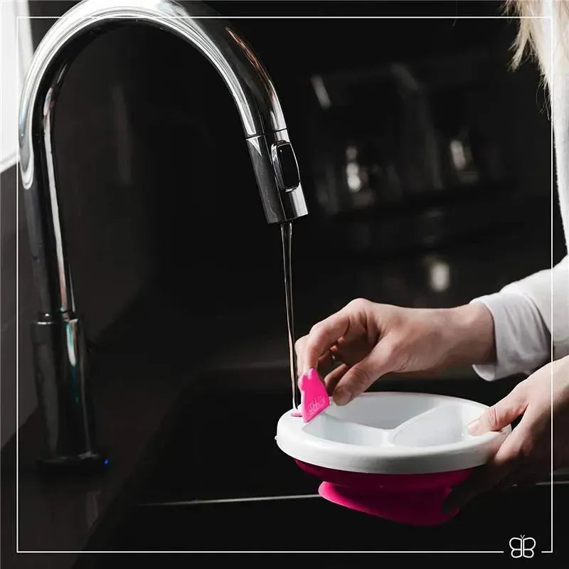 Hands holding a Bbluv - Platö warm feeding plate in pink under running water from a stylish faucet.