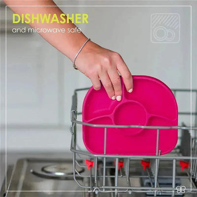 A hand placing a pink Bbluv Yümi 4-section silicone plate in a dishwasher, labeled as dishwasher and microwave safe.