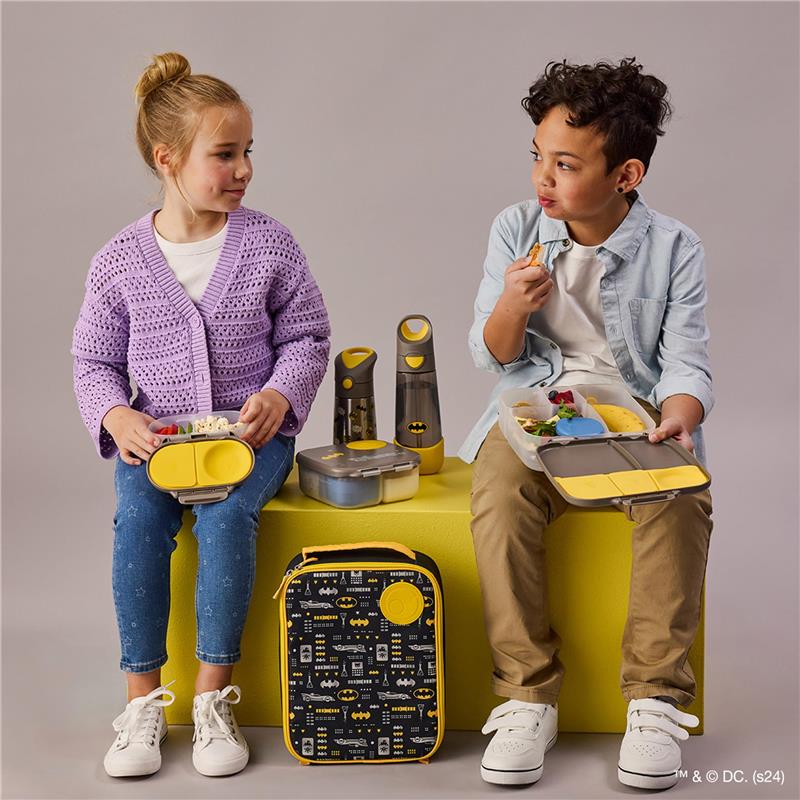 Two children sit on a yellow stool with a Batman-themed lunch bag and water bottles. The girl holds a lunchbox with snacks, while the boy enjoys food from his container.
