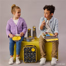 Two children sit on a yellow stool with a Batman-themed lunch bag and water bottles. The girl holds a lunchbox with snacks, while the boy enjoys food from his container.