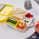 Lunchbox with compartments containing sandwiches, cucumber slices, an apple, yogurt with blueberries, and strawberries, beside a drink.