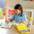A girl in a blue shirt interacts with a Bbox Lunchbox Emerald Forest, featuring fruit and sandwiches, while working on her homework.
