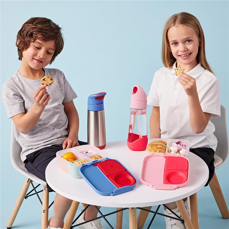 Two kids sitting at a table with a blue and red Bbox lunch box, snacks, and water bottles, smiling and enjoying their food.