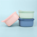 Silicone lunch tubs in pink, green, and blue stacked on a light blue background.