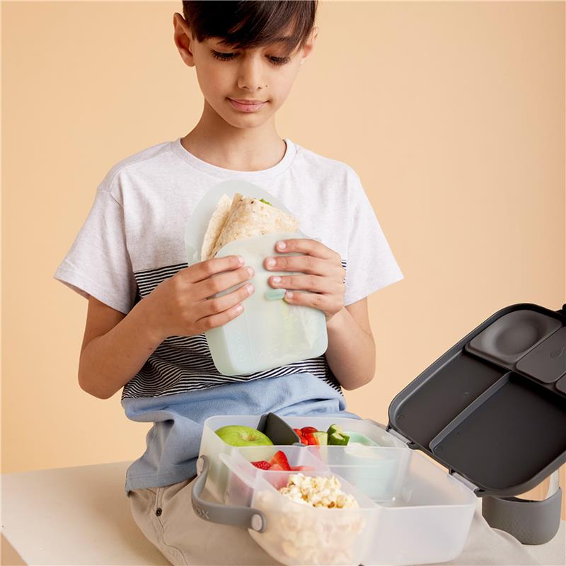A boy holds a silicone sandwich pocket filled with a wrap, beside a multi-compartment lunchbox with fruits, vegetables, and popcorn.