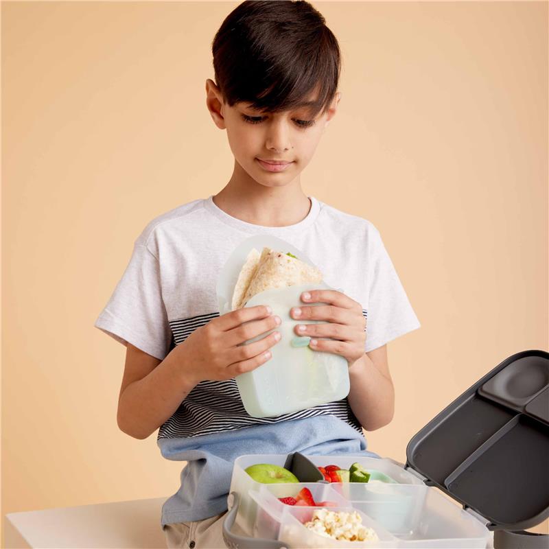 A young boy holds a silicone sandwich pocket filled with wraps, beside a lunchbox containing fruits and snacks.
