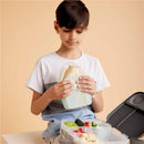A young boy holds a silicone sandwich pocket filled with wraps, beside a lunchbox containing fruits and snacks.