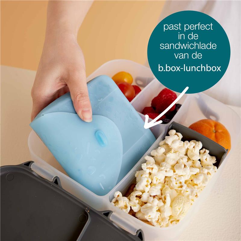 Silicone sandwich pocket in blue being placed in a b.box lunchbox with popcorn, fruit, and berries visible.