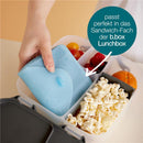 Silicone sandwich pocket in light blue being placed in a b.box lunchbox, with popcorn, fruits, and text indicating its purpose.