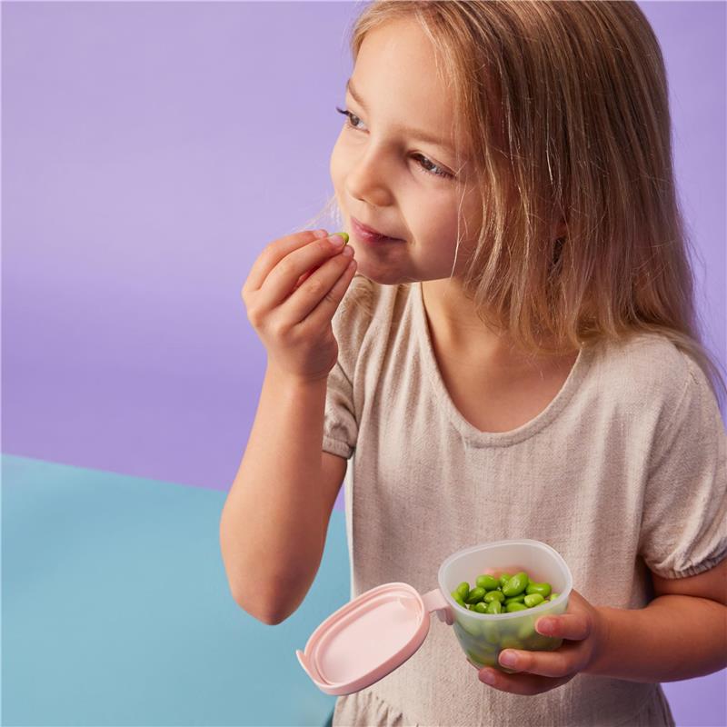 Child enjoying green snacks from a pink Bbox silicone snack tub against a colorful background.