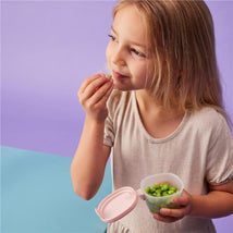 Child enjoying green snacks from a pink Bbox silicone snack tub against a colorful background.