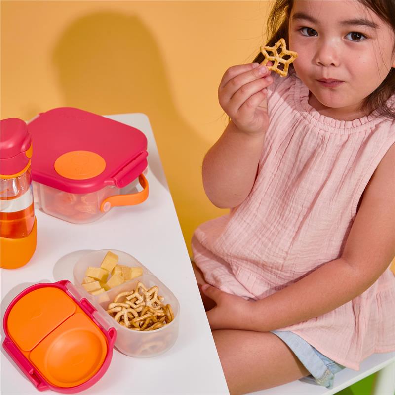 A young girl in a pink top holds a star-shaped snack, with a colorful snack box and drink on the table.