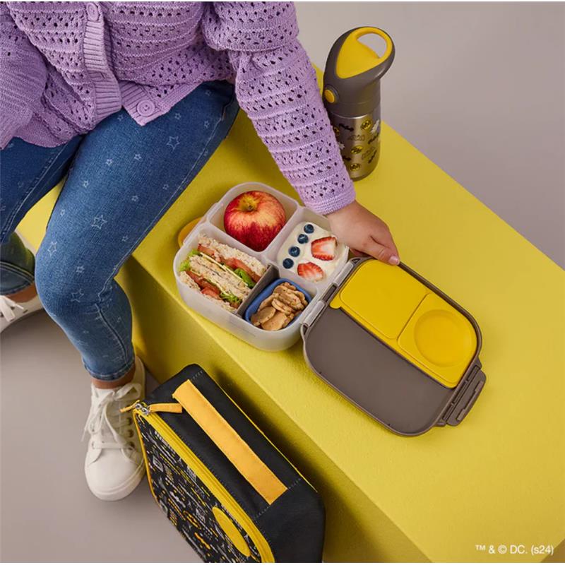 Batman-themed mini lunch box with sandwich, apple, yogurt, and snacks, paired with a water bottle, on a yellow surface.