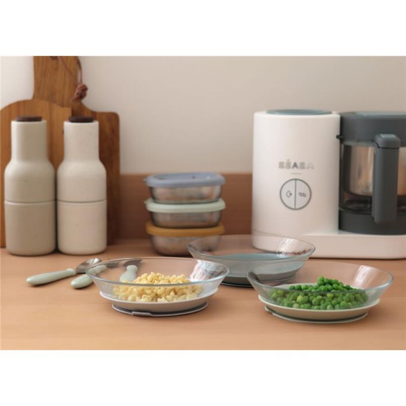 Beaba 3Pk Glass Plate Set in Grey Green with three plates holding cooked grains and peas, alongside kitchen utensils and a food processor.