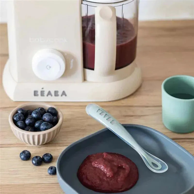 Beaba Babycook Solo 4 in 1 Baby Food Maker in Oat color, with pureed food, blueberries, and a spoon on a wooden surface.