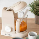 Beaba Babycook Solo 4 in 1 Baby Food Maker in oat color, with a transparent bowl containing orange puree and a spatula.