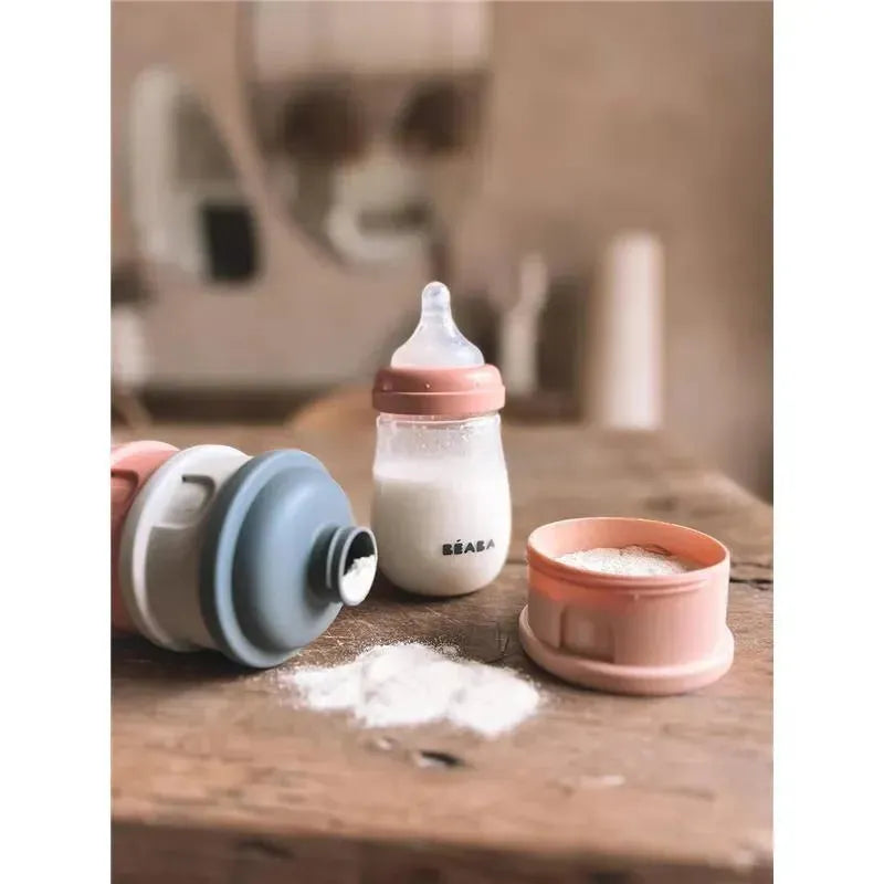 Beaba formula and snack container in rose color with a milk bottle and powdered formula on a wooden surface.