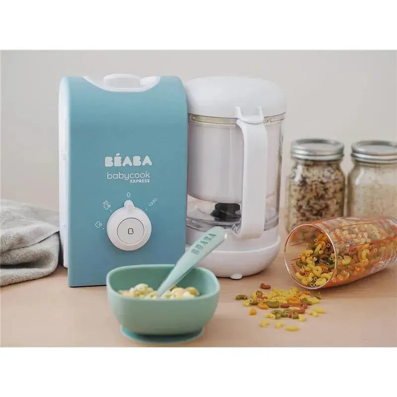 Beaba Babycook Express in light blue, a bowl of cooked grains, a measuring spoon, and colorful pasta scattered nearby.