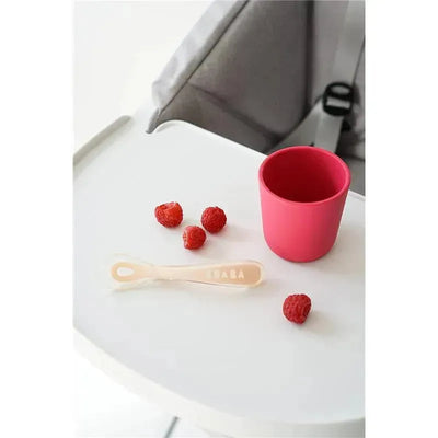 Self-feeding silicone spoon from Beaba in pink, placed on a high chair tray with raspberries and a matching cup.
