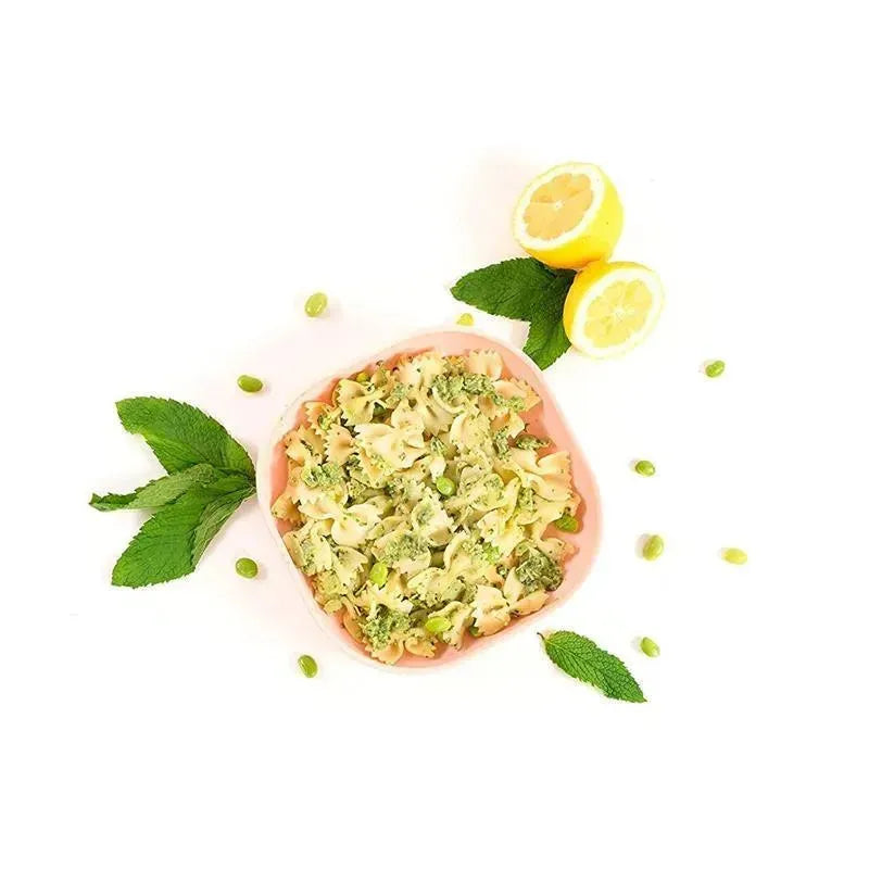 Silicone meal set featuring a pink plate filled with pasta mixed with broccoli and edamame, garnished with lemon and mint leaves.