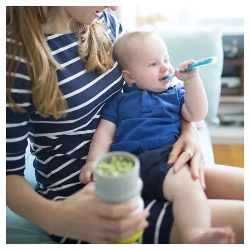 Mom feeding a baby with a Beaba silicone spoon while holding a food container; both are seated on a couch.