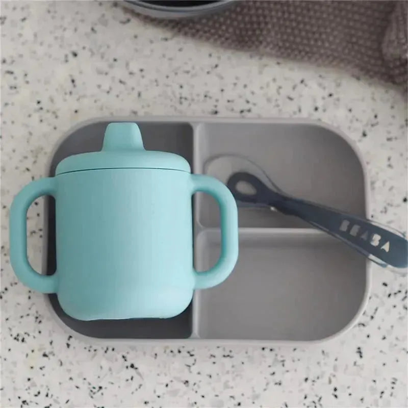Silicone training cup by Beaba in light blue with two handles, placed on a gray divided tray alongside a spoon.