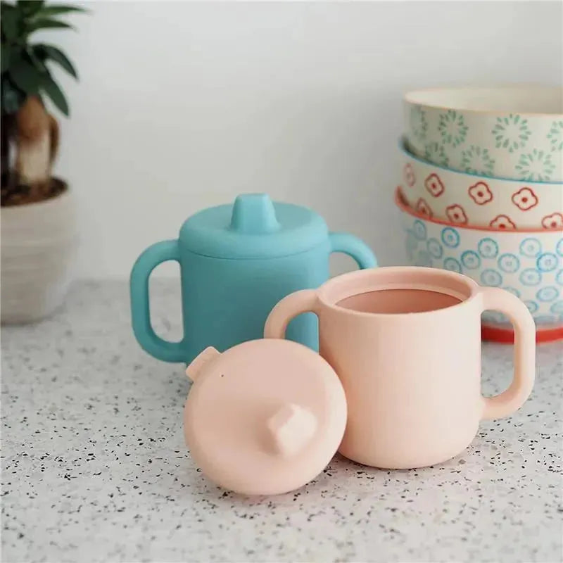 Silicone training cup set by Beaba in pink and blue, featuring ergonomic handles and a matching lid, on a speckled surface.