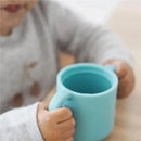 Silicone training cup by Beaba in teal, held by a child, designed for easy gripping during drinking.