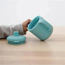 Silicone training cup in rain color being held, with a matching lid placed nearby on a wooden surface.