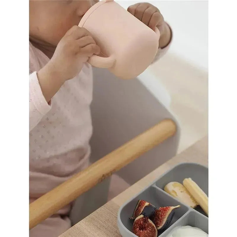 Silicone training cup in rose color held by a child, with fruits and yogurt on a food tray nearby.