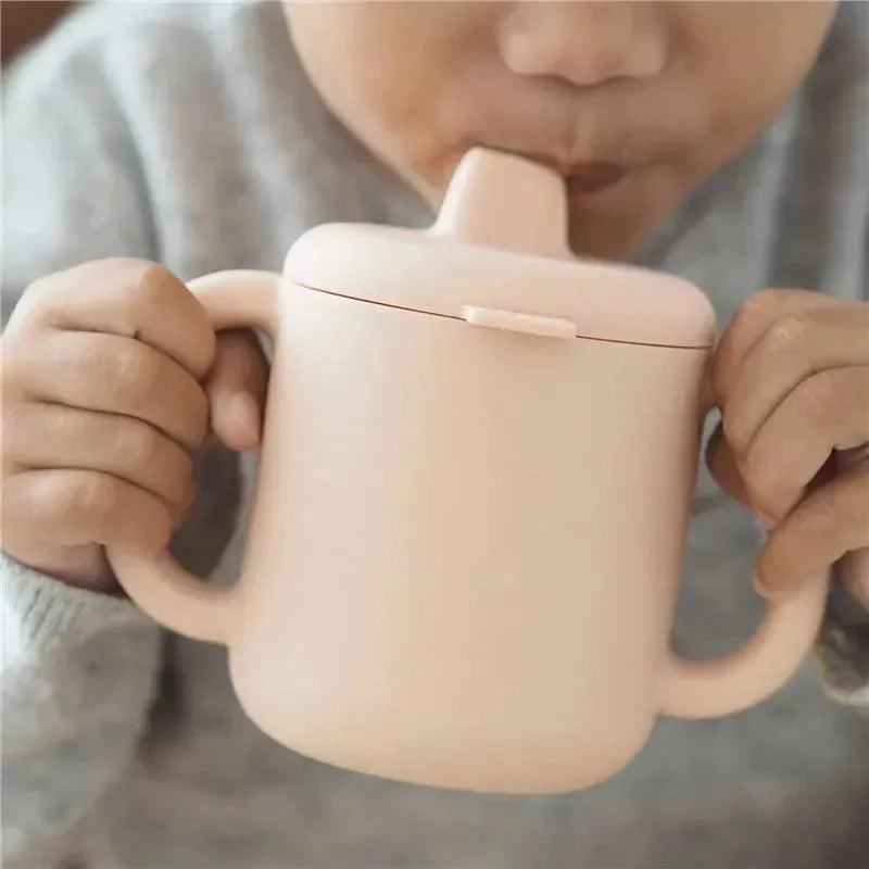 A child holds a soft pink Beaba silicone training cup with a spout, designed for easy drinking.