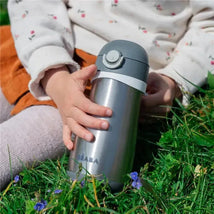 Stainless steel kids water bottle from Beaba in charcoal, held by a child in a grassy area with small flowers.