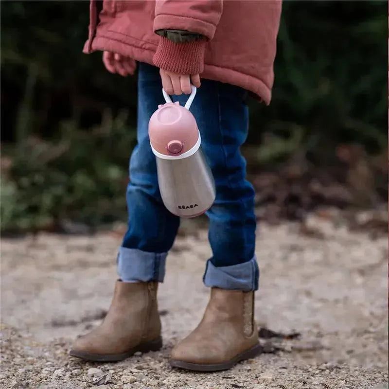 Stainless steel kids water bottle by Beaba in rose color, held by a child wearing a pink jacket and brown boots.