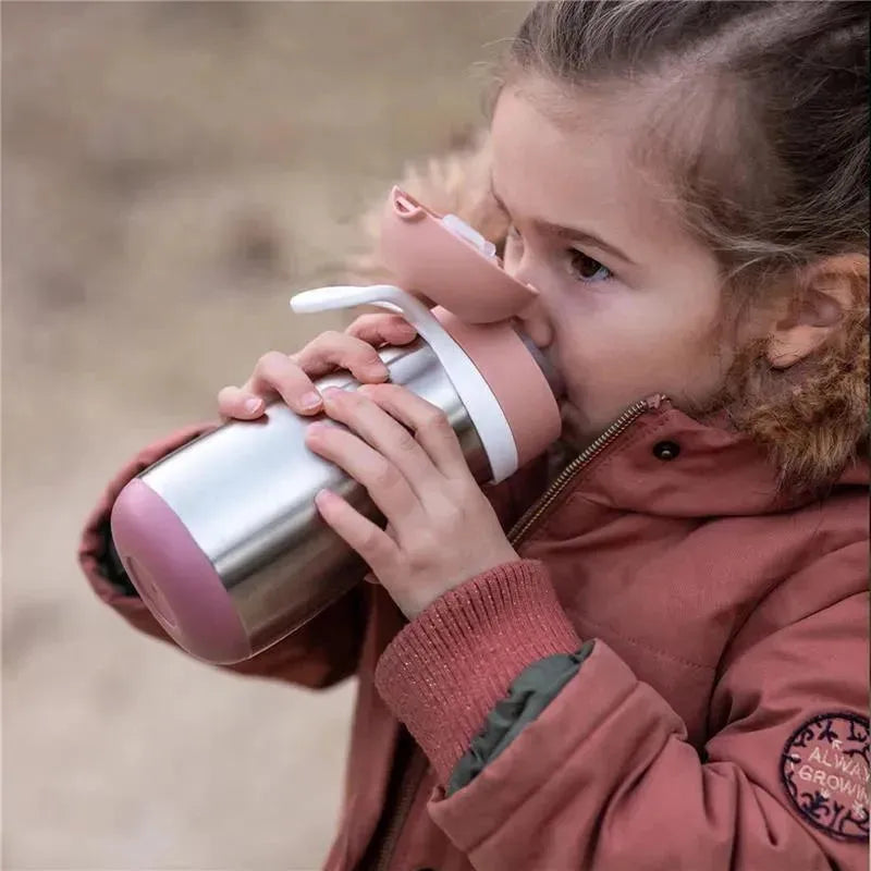 Child drinking from a Beaba stainless steel kids water bottle in rose color, with a soft pink lid and a straw.