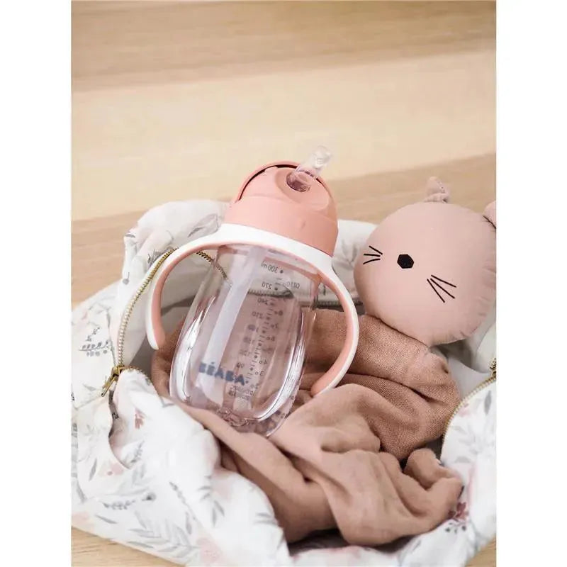 Beaba rose straw sippy cup with clear body and handles, placed beside a cuddly cat toy in a soft fabric bag.