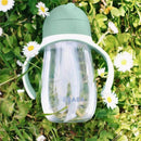 Beaba straw sippy cup in sage green color, with clear body and handles, surrounded by small white daisies on grass.