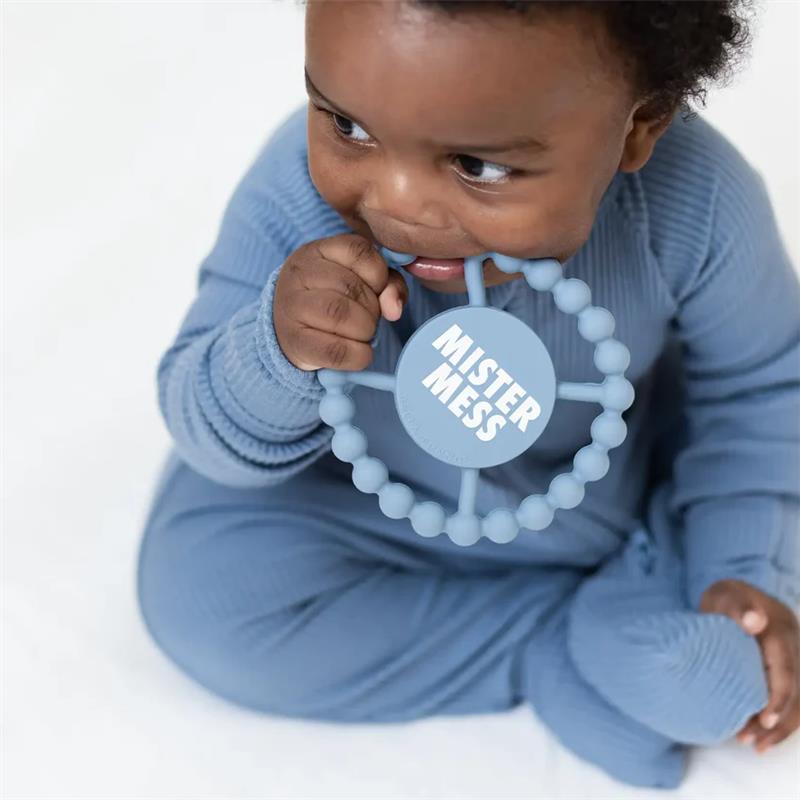 A baby in a blue outfit bites the Bella Tunno Mister Mess teether, featuring a circular design with beads and text.
