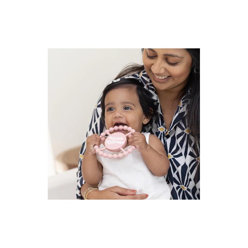 Infant holding a Bella Tunno Happy Teether with a pink beaded design, smiling at the caregiver.