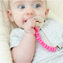 Baby holding a pink Bella Tunno Punch Pacifier Clip attached to a pacifier, sitting in a crib.