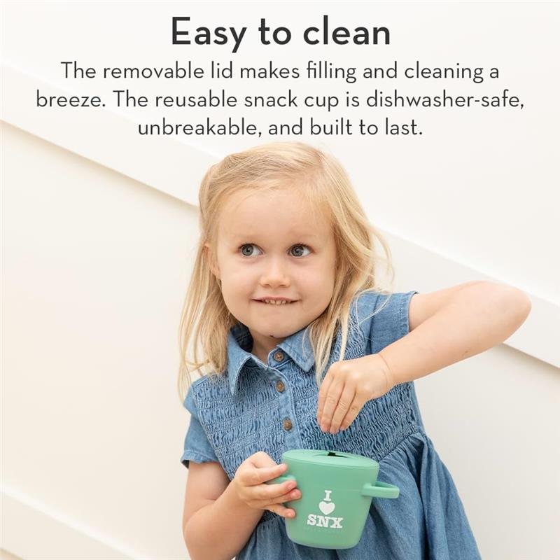 A young girl in a denim dress holds a green silicone snack cup with a lid, smiling while reaching inside.