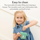 A young girl in a denim dress holds a green silicone snack cup with a lid, smiling while reaching inside.